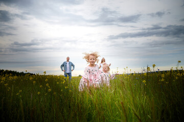 A family including two young parents and daughters on a walk in a meadow with grass and flowers. Dad, mom, girls relaxing and having fun in nature on a summer day with clouds