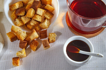 Square toasted pieces of homemade delicious rusk, hardtack, Dryasdust, zwieback, Liquid honey in a saucer and black tee in a cap on a white tablecloth.