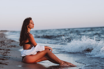 Fototapeta na wymiar A young beautiful woman in a black bikini and a white shirt on a tanned body sits on the beach in the waves. Soft selective focus.