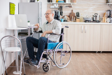 Senior man in wheelchair using laptop in the kitchen. Disabled senior man in wheelchair having a video conference on laptop in kitchen. Paralyzed old man and his wife having a online conference.