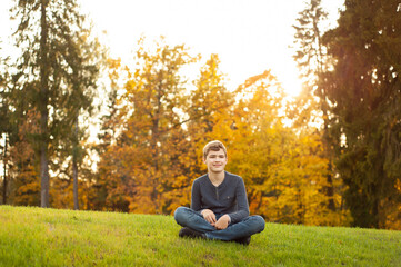 Fototapeta premium A teenage boy sits on the grass in an autumn park. Autumn walk.