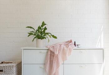 Close up of white dresser against painted brick wall with pot plant, pink scarf and lotions...
