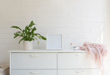 Close up of white dresser against painted brick wall with pot plant, blank square frame, pink scarf...