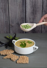 Soup-mashed peas and beans in a white Cup on a gray wooden background with grain crackers, hand with a spoon with peas over a Cup of soup . Place for a copy space