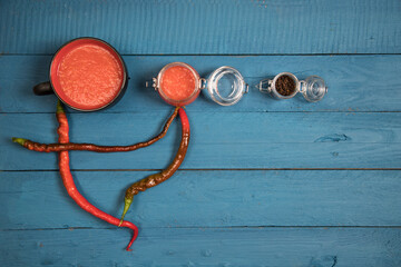 Hot red pepper sauce on a wooden blue background in a glass jar and Cup, red capsicum lies next to it. Place for a copy space