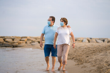 pensioner woman and her husband taking romantic walk together - happy retired mature couple in face mask walking on the beach in new normal holidays trip during covid19