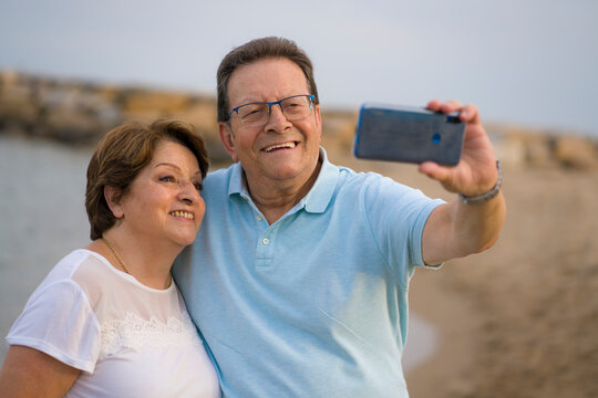 happy pensioner woman and her husband taking romantic walk taking selfie - happy retired mature couple walking on the beach during holidays taking self portrait with mobile phone