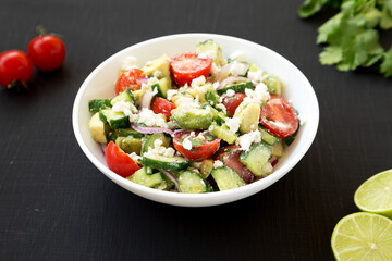 Delicious Avocado Tomato and Cucumber Salad in a white bowl on a black surface, low angle view. Close-up.