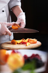 The cook cuts the fruit into slices. Assorted fruits and berries are cut into pieces. Unrecognizable vertical photo. Only hands with gloves.