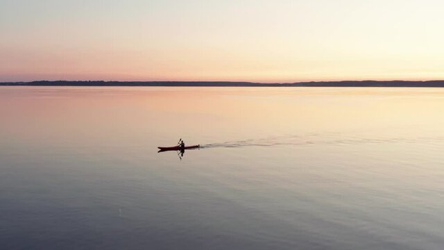 Sunset paddle in drone shot. Aerial shot of canoe or kayak paddling on windless calm water in red orange purple evening light. Sport activity on lake or sea in beautiful healthy natural environment