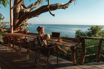 Fototapeta Pretty young woman using laptop in cafe with sea view. Freelancer working remotely from beach cafe, drink coconut and use laptop obraz