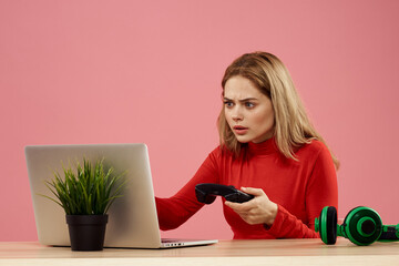 Woman sitting at a table in front of a laptop with a gamepad in hands playing technology headphones 