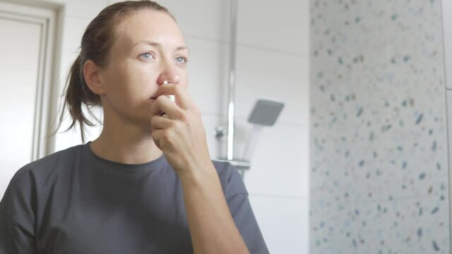A woman throws drops in the nose of the house in front of the mirror. Reflection in the bathroom mirror.