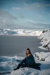 Fototapeta na wymiar Lifestyle portrait of young woman sitting next to a frozen lake in snow. Cuddled up in a blanket she is posing for a camera.