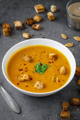 Pumpkin soup with coriander, gluten free croutons, chili and garlic flakes seasoning in a white bowl. Croutons, pumpkin seeds and a candle in the background. Seasonal autumn / winter food.