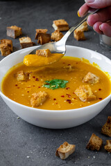 Woman hand taking a spoon of pumpkin soup from a white bowl with coriander, gluten free croutons and chili / garlic flakes seasoning. More crouton in the background. Autumn Winter food
