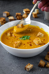 Woman hand taking a spoon of pumpkin soup from a white bowl with coriander, gluten free croutons and chili / garlic flakes seasoning. More crouton in the background. Autumn Winter food