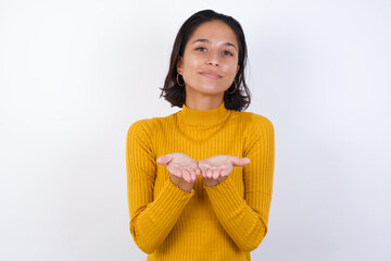 Young hispanic girl with short hair wearing casual yellow sweater isolated over white background holding something with open palms, offering to the camera.