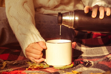 girl in warm white sweater on plaid blanket pours coffee from thermos. Autumn background, cncept of warmth and comfort