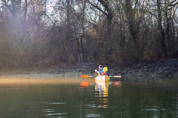 Fototapeta na wymiar Kayaking in winter at sunrise in a foggy haze in the Danube river. A man and a woman row in a two-person kayak