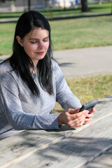 Attractive young woman sitting at a picnic table using her smart phone to check her messages in the park.