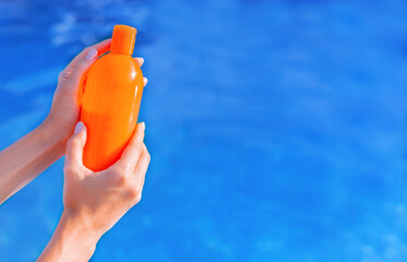 Orange bottle of sun protection body suntan lotion, moisturizing sunblock cosmetic sunscreen product in female hand against swimming pool in summer time. Skin body care protection Copy space