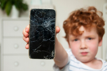 Close-up of young boy with serious and worried expression holding a broken smart phone in a home setting. Focus on phone, with boy blurred.