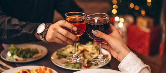 hands with glasses clinking against the background of Christmas tree lights and bonfires from a home fireplace over a table with delicious dishes