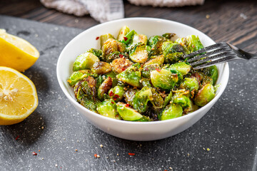 Roasted / fried brussels sprouts in a white bowl sitting on a cutting board with a fork ready for eating. Lemon halves, towel, chili and garlic flakes seasoning in the background	