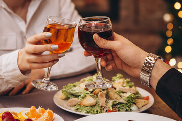 hands with glasses clinking against the background of Christmas tree lights and bonfires from a home fireplace over a table with delicious dishes