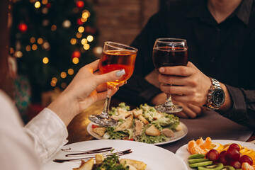 close up hands with glasses clinking against the background of Christmas tree lights and bonfires from a home fireplace over a table with delicious dishes