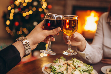 close up hands with glasses clinking against the background of Christmas tree lights and bonfires from a home fireplace over a table with delicious dishes