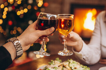 close up hands with glasses clinking against the background of Christmas tree lights and bonfires from a home fireplace over a table with delicious dishes