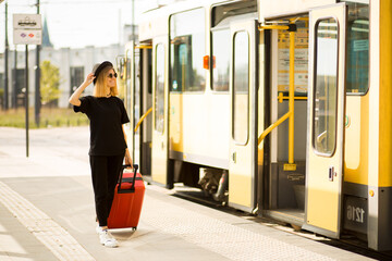 Stylish woman wear total black with red travel suitcase stands at stop awaiting tram - obrazy, fototapety, plakaty