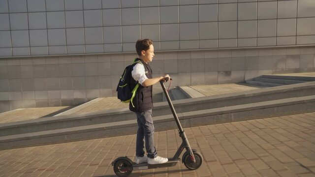 School boy riding with his electric scooter in the city with backpack on sunny day. Child in colorful clothes biking on way to school. Electric transport.