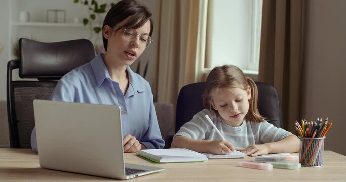 Portrait of two female persons, young smiling woman uses computer for presentation in lesson with little cute preschool girl, shows her video or photo, teaches to write and read, sit together at table