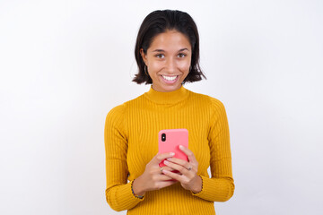 Young hispanic girl with short hair wearing casual yellow sweater  enjoys distant communication, uses mobile phone, surfs fast unlimited internet, has pleasant smile, makes shopping online, 