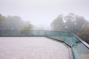 Autumn fog in the old majestic park. Colorful leaves on the trees. Glass panels on the edge of the terrace. Outdoor summer terrace.