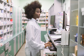 Side view portrait of young confident concentrated African female pharmacist, working with computer...
