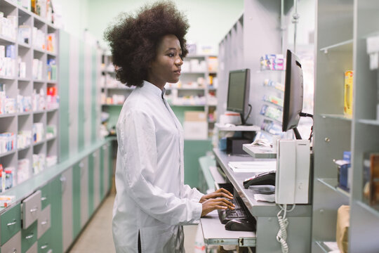 Side view portrait of young confident concentrated African female pharmacist, working with computer behind counter in pharmacy