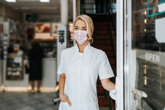 Young and attractive female pharmacist with face protective mask standing at open drugstore doors and looking outside. She is confident and serious. Covid-19 open for business concept.