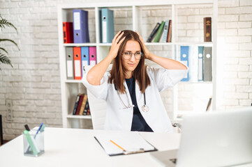 Upset young female doctor looking at laptop and holding head with hands reading bad news online. Young woman physician has problem at work