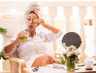 Beautiful woman wearing white bathrobe with cucumber skin care mask while drinking detox drink. DIY...