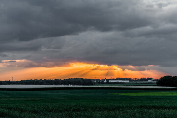 Sunset in a panoramic landscape of a green field cultivated with the rays of the sun rising through the clouds on a cloudy day in Beek, South Limburg in the Netherlands