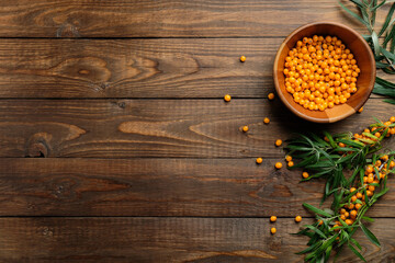 Sea buckthorn berries in bowl with plant branches on wooden desk. Flat lay, top view, copy space.