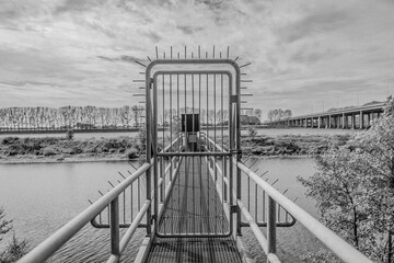 Metal bridge with a closed door prohibiting passage, bare trees and road bridge A76 (Scharbergbrug) over the Maas river in the background, South-Limburg in the Netherlands. Black and white image