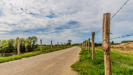 Rural road between fences with wooden posts and barbed wire in the middle of a Dutch countryside, sunny day with blue sky with white clouds between Elsloo and Meers, South-Limburg in the Netherlands
