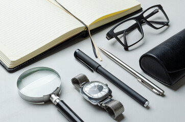 Closeup of magnifying glass, male wathces, pens, glasses, case and opened notebook on the desk
