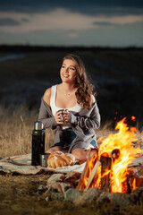 Beautiful girl drinking tea by the fire. Pcnic rest. Thermos tea. Romantic atmosphere in nature. Campfire. Bonfire