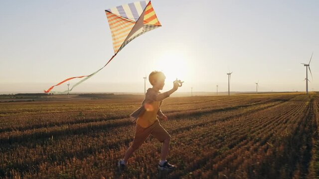 Happy boy runs launches bright kite into sky mown wheat field, playing with wind in field of an orange sunset on day lens flares wind turbines in summer slow motion. School break. Lifestyle. Childhood
